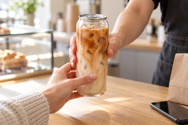 A barista hands a sealed can of iced latte to a customer across the counter.